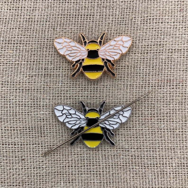 May include: Two gold bee pins with black and yellow stripes and white wings. One bee pin is on a beige fabric surface and the other bee pin is on a needle.