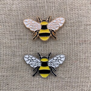 May include: Two gold-colored enamel pins shaped like bumblebees. The bees have black and yellow stripes, white wings, and black antennae.