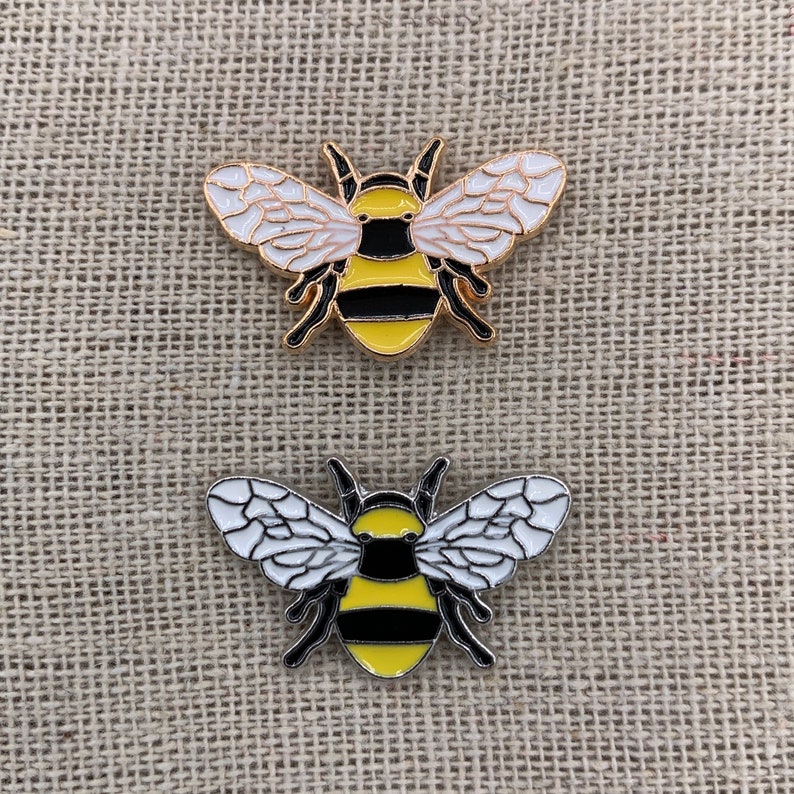 May include: Two gold-colored enamel pins shaped like bumblebees. The bees have black and yellow stripes, white wings, and black antennae.