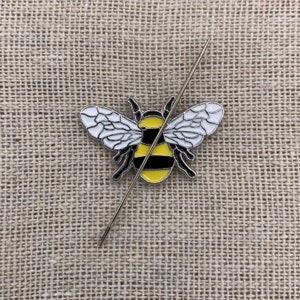 May include: A yellow and black bumblebee pin with white wings, pinned to a beige fabric background with a silver needle.