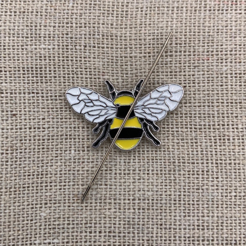 May include: A yellow and black bumblebee pin with white wings, pinned to a beige fabric background with a silver needle.