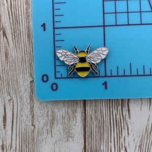 May include: A yellow and black enamel bee pin with white wings. The pin is on a blue background with a ruler.