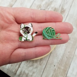 K&ouml;nnte beinhalten: Zwei Emaille-Pins, einer mit einer wei&szlig;en Katze mit gr&uuml;nem Garn und der andere mit einer gr&uuml;nen Wollkn&auml;uel.