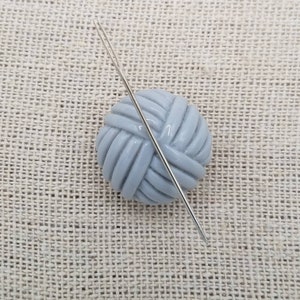 May include: A gray ceramic yarn ball with a silver needle sticking through it. The yarn ball is on a beige textured surface.