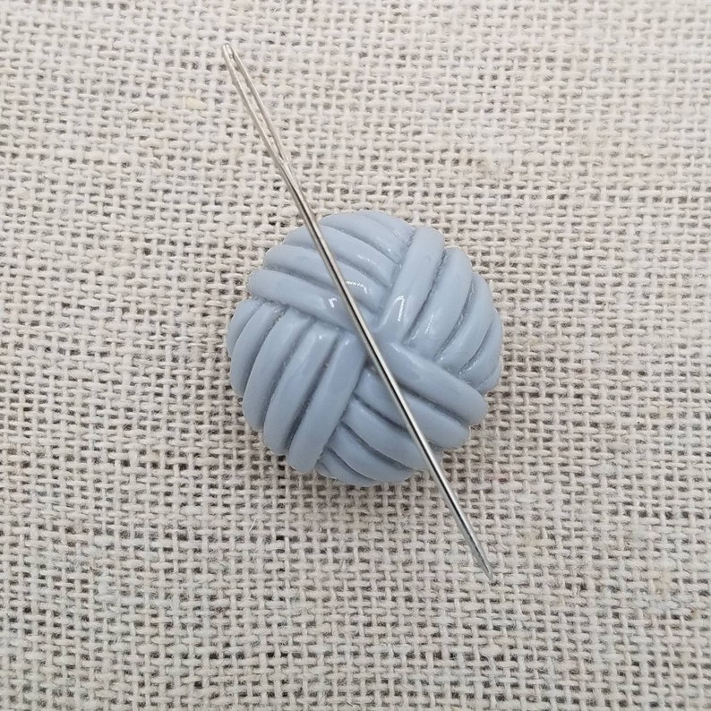 May include: A gray ceramic yarn ball with a silver needle sticking through it. The yarn ball is on a beige textured surface.