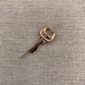 May include: A gold-colored key is pierced by a thin, silver needle on a beige fabric background.