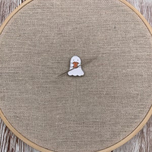 Peut inclure: Un fant&ocirc;me blanc tenant une petite citrouille orange, avec une aiguille et du fil passant &agrave; travers le fant&ocirc;me, sur un fond de toile de jute brune.