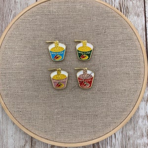 May include: Four enamel pins shaped like a bowl of noodles with a red chilli pepper. The pins are in different colours: blue, green, pink, and red. The pins say "NOODLES" on the front.