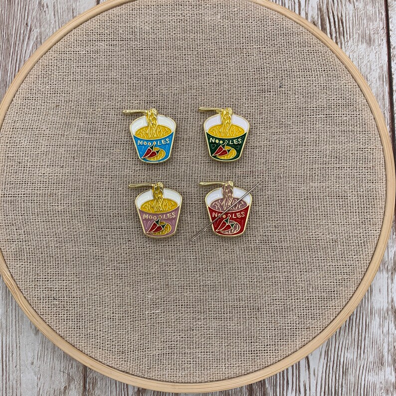 May include: Four enamel pins shaped like a bowl of noodles with a red chilli pepper. The pins are in different colours: blue, green, pink, and red. The pins say "NOODLES" on the front.