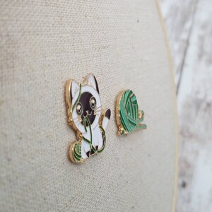 K&ouml;nnte beinhalten: Zwei Emaille-Pins mit einem wei&szlig;en K&auml;tzchen mit schwarzen Markierungen, das mit einem gr&uuml;nen Wollkn&auml;uel spielt. Die Katze schaut das Garn mit einem verspielten Ausdruck an.