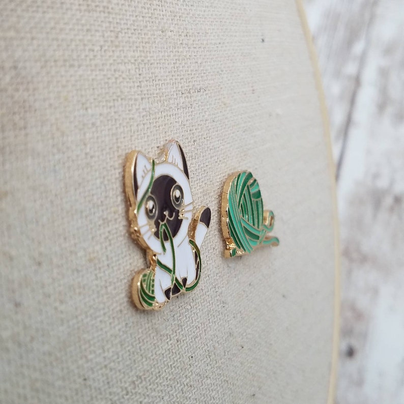 K&ouml;nnte beinhalten: Zwei Emaille-Pins mit einem wei&szlig;en K&auml;tzchen mit schwarzen Markierungen, das mit einem gr&uuml;nen Wollkn&auml;uel spielt. Die Katze schaut das Garn mit einem verspielten Ausdruck an.