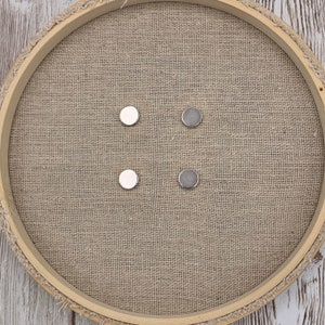May include: Four round magnetic buttons, two silver and two white, arranged in a square pattern on a brown hessian surface.