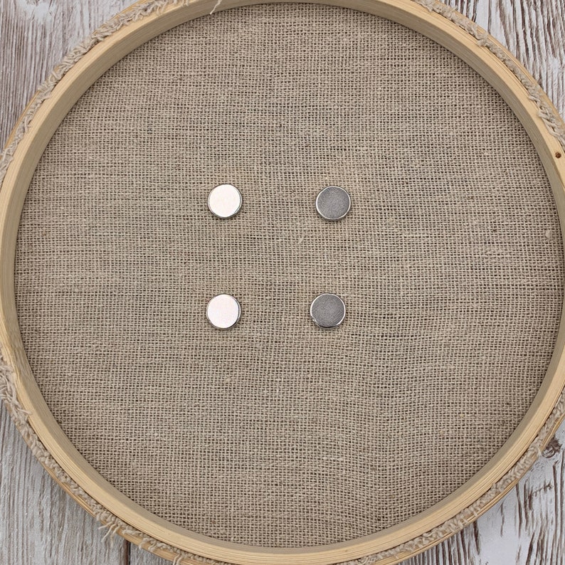 May include: Four round magnetic buttons, two silver and two white, arranged in a square pattern on a brown hessian surface.