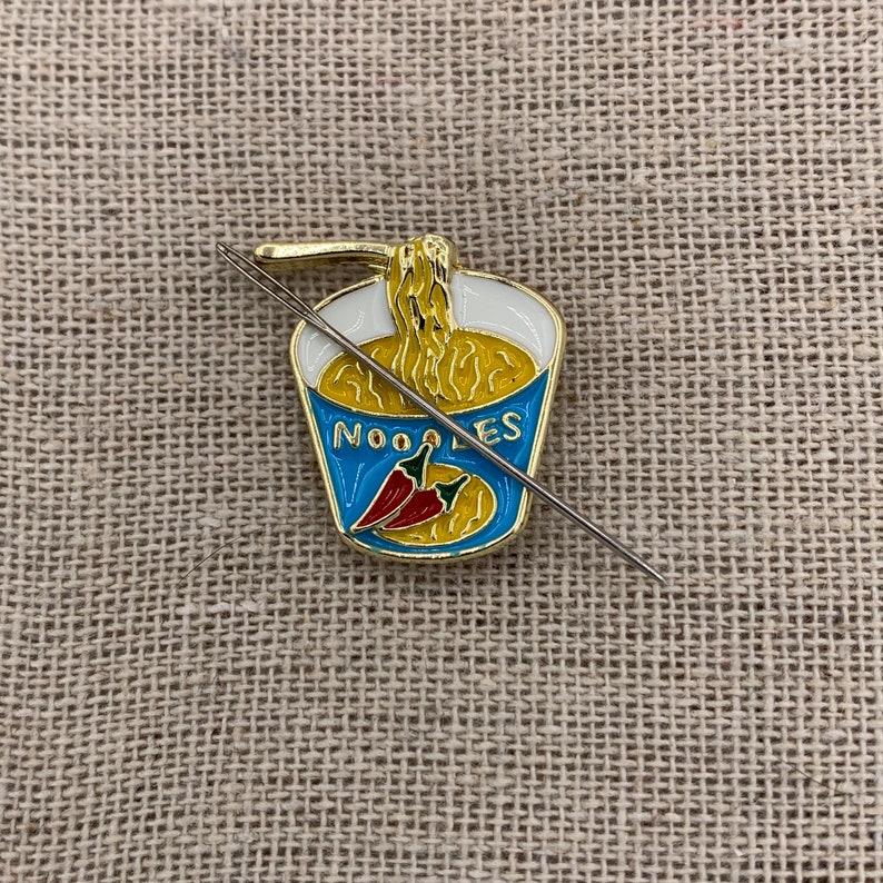 May include: A gold-coloured enamel pin shaped like a cup of noodles with a red chilli pepper and the word "NOODLES" on the front. A silver needle is threaded through the pin.