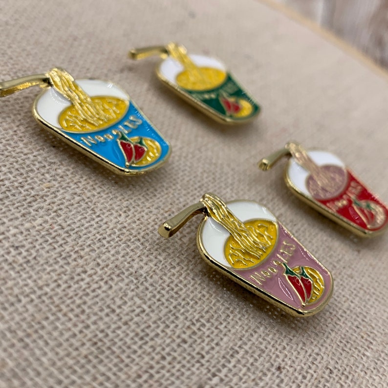 May include: Four enamel pins shaped like a cup of noodles with a straw. The pins are in different colours: blue, green, pink, and red. Each pin has a red chilli pepper and the text "Noodles" on it.
