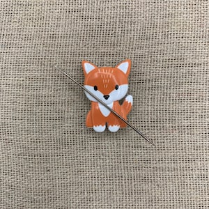 May include: A small orange fox-shaped needle minder with a white face and a silver needle threaded through it. The needle minder is on a brown burlap background.