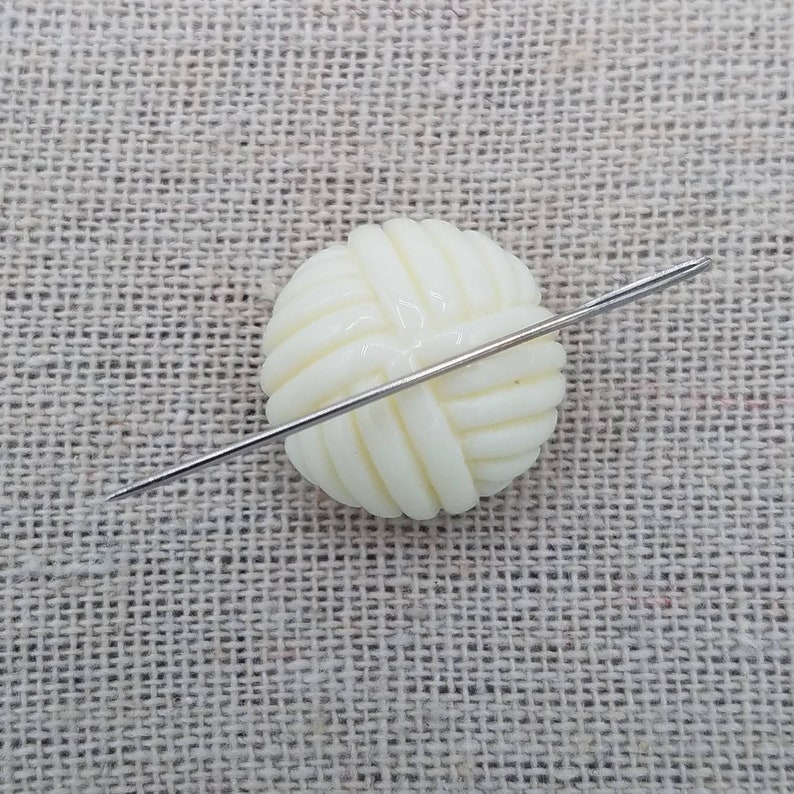 May include: A white plastic button shaped like a ball of yarn with a silver needle going through it.