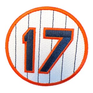 May include: A white and orange baseball jersey patch with the number 17 in black.