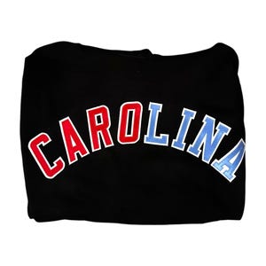 Puede incluir: Sudadera con capucha negra con un dise&ntilde;o de arco "CAROLINA" en rojo y azul.