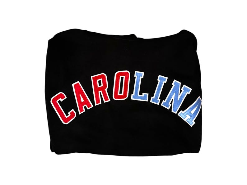Puede incluir: Sudadera con capucha negra con un dise&ntilde;o de arco "CAROLINA" en rojo y azul.
