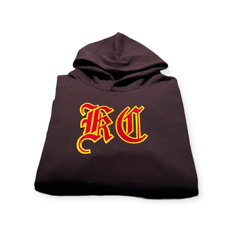 May include: A dark brown hooded sweatshirt with a red and yellow embroidered "KC" logo on the front.
