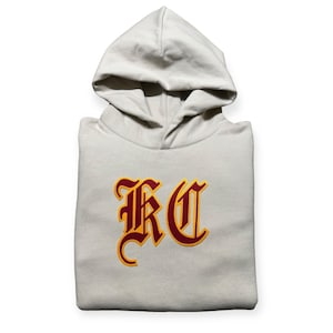 May include: A beige hooded sweatshirt with a large red and yellow "KC" logo on the front.
