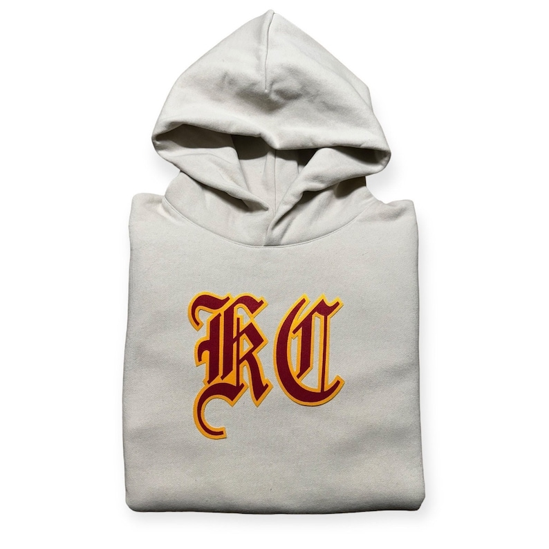 May include: A beige hooded sweatshirt with a large red and yellow "KC" logo on the front.