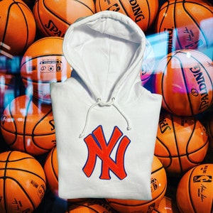 May include: A white hoodie with a drawstring hood and a large orange and blue "NY" logo on the front. The hoodie is displayed against a backdrop of orange basketballs.