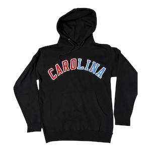 Puede incluir: Sudadera con capucha negra con la palabra "CAROLINA" en letras rojas, blancas y azules en forma de arco.