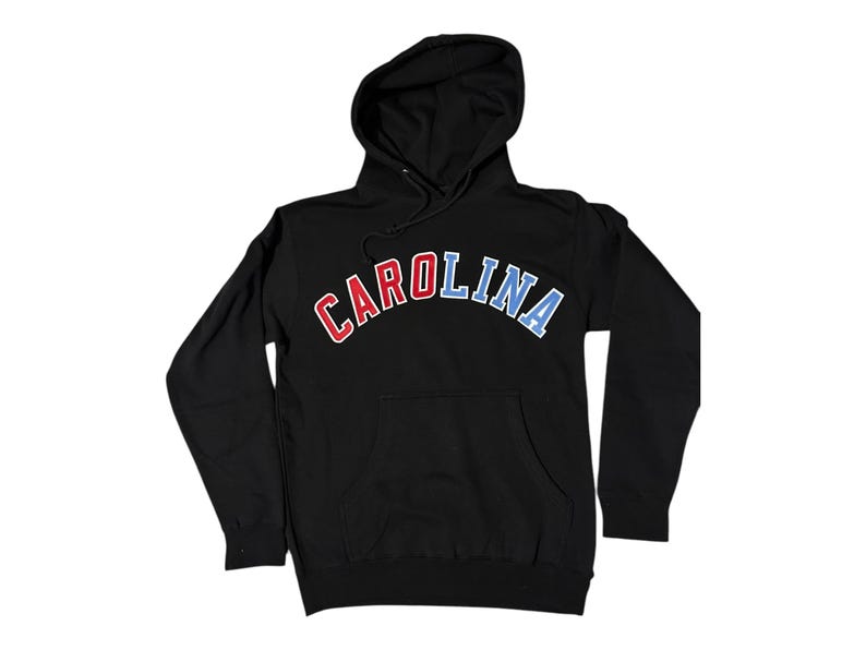Puede incluir: Sudadera con capucha negra con la palabra "CAROLINA" en letras rojas, blancas y azules en forma de arco.