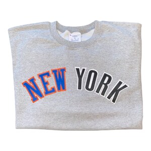 May include: Gray Champion sweatshirt with a large, arched "NEW YORK" graphic in blue, orange, and black lettering.