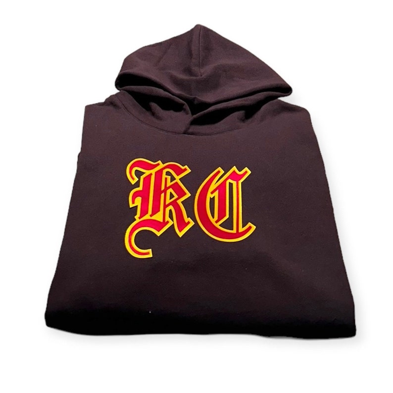 May include: A dark brown hooded sweatshirt with a red and yellow "RC" logo in a gothic font.