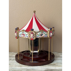 May include: A carousel-style display stand featuring a red and white striped canopy, gold-toned twisted support poles, and decorative panels with sculpted clown faces. The base is made of dark wood with circular indentations for storage.