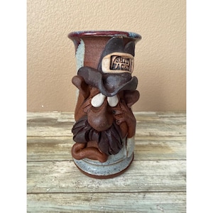 May include: A handmade ceramic vase with a sculpted face and hat. The vase is primarily brown with accents of blue, red, and white. The hat has the text "SHIT FACE" on it. The vase has a rustic, folk art style.