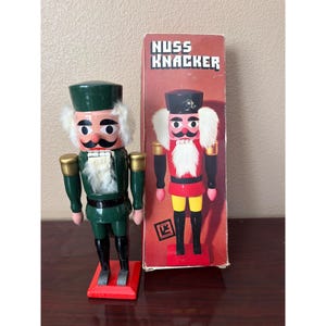 Puede incluir: Una figura de cascanueces de madera pintada en verde, negro y dorado con pelo y barba de piel sintética blanca, junto a su caja de cartón roja original. La caja presenta el texto NUSS KNACKER y una ilustración de un cascanueces rojo.