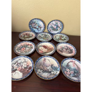 May include: A collection of ten round decorative plates featuring various floral arrangements in woven baskets. Each plate has a dark blue border with a repeating white and gold floral pattern and a thin gold rim around the outer edge.