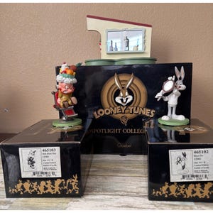 May include: A collection of Looney Tunes figurines, including Bugs Bunny and Yosemite Sam, displayed with their original boxes. The figurines are set against a backdrop featuring a Looney Tunes logo and a miniature barbershop scene.