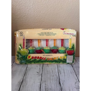 Könnte beinhalten: Eine Vintage-Box mit 7 Classic Bubble Light Set. Die Box hat ein klares Fenster, das die bunten Lichter zeigt. Jede Leuchte hat eine rote, grüne oder gelbe Basis mit einem transparenten Blasentop. Auf der Box steht "Classic" und "See Them Bubble!"