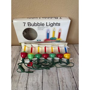 Puede incluir: Una caja de 7 luces de burbujas con un diseño retro. Las luces tienen bombillas rojas, verdes y amarillas con un cable verde. La caja tiene el texto "7 Bubble Lights".