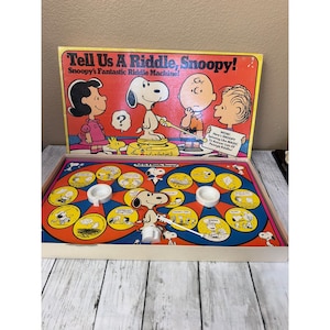 May include: Vintage Snoopy board game, "Tell Us A Riddle, Snoopy!" The box features Snoopy, Lucy, and Charlie Brown. The game board is orange and blue with Snoopy and other Peanuts characters. The box is open, revealing the game pieces.