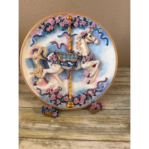 May include: A round decorative plate featuring a three-dimensional white carousel horse with a blue and green saddle. The horse is surrounded by sculpted pink roses and blue ribbons against a soft blue and pink background with a gold rim.