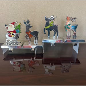 May include: A set of four silver-toned metal stocking holders featuring holiday designs. The figures include Santa Claus in a sleigh with a tree, and three reindeer wearing scarves in red, green, and teal colours, all mounted on rectangular bases.