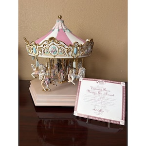 May include: A Victoria Rose Merry-Go-Round carousel figurine featuring a pink and white striped canopy with gold trim, blue accents, and white horses. A certificate of authenticity is displayed in front, titled Victoria Rose Merry-Go-Round.