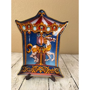 May include: A decorative tray with a flared, pagoda-like shape featuring a carousel horse illustration. The design includes a white horse with gold accents, a blue background with stars, and ornate gold scrollwork borders with red and white stripes.