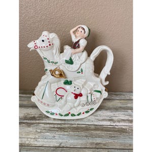May include: A white ceramic teapot shaped like a rocking horse with a child rider. The design features red and green holly accents, a gold-coloured horn, a teddy bear, a toy sailboat, and a train engine. The handle is curved and integrated.