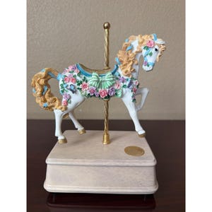 May include: A white carousel horse figurine with a flowing blonde mane and tail, adorned with pink and purple floral garlands, a green bow, and blue butterflies. The horse is mounted on a gold-colored metal pole atop a beige rectangular base.