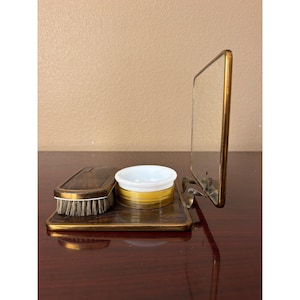May include: A vanity set featuring a rectangular base with a dark wood-grain finish and a brass border. It includes a small brush with a dark patterned top, a white and yellow circular container, and an upright rectangular mirror with a brass frame.