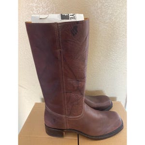 May include: A pair of brown leather riding boots. The boots are tall, reaching the mid-calf, with a rounded toe and a low heel. The leather has a distressed look, with visible creases and variations in colour. The Frye logo is visible on the side.