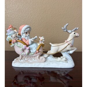 Könnte beinhalten: Eine Porzellanfigur, die eine als Weihnachtsmann gekleidete Figur in einem rosa Schlitten zeigt, der mit Geschenken, einem Teddybären und einem kleinen Tier gefüllt ist. Ein weißes Rentier mit Goldketten ist am Schlitten befestigt, mit einem Hasen.