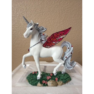 May include: A white unicorn figurine with a silver horn, mane, and tail. The unicorn features large wings with a red and pink stained-glass pattern, a silver chain necklace, and small red gems embedded in its mane, tail, and wings. It stands on a green base.
